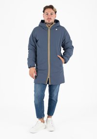 Mannen lange, marineblauwe jas met capuchon, gele ritsaccent en twee zijzakken. G gedragen over blauwe jeans en witte sneakers.
