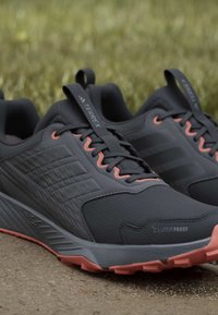 Chaussures de trail running gris foncé et corail avec semelles texturées antidérapantes, de marque "Terrex" et "Climaproof", posées sur une surface pavée avec un fond de gazon.