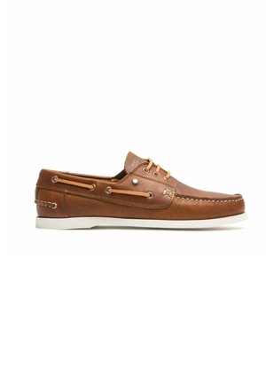 Des chaussures bateau en cuir brun avec des semelles en caoutchouc blanc, présentant des œillets en métal et des lacets beige. Des coutures sur le côté ajoutent un détail.