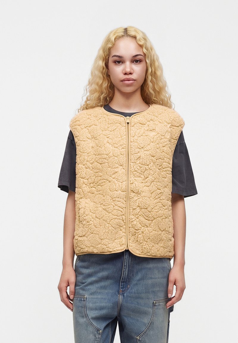 Jeune personne aux longs cheveux blonds bouclés portant un gilet zippé beige texturé par-dessus un t-shirt sombre à manches courtes et un jean bleu.