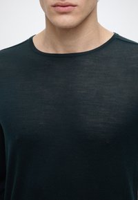 Jeune personne portant un pull vert foncé à col rond, visible du cou jusqu'à la partie supérieure de la poitrine sur un fond clair uni.
