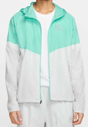 Mężczyzna w jasnoturkusowej i białej kurtce Nike Air z zamkiem, białej podkoszulce i dopasowanych białych spodniach z logo.