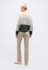 Άτομο με κοντά ξανθά μαλλιά, φορώντας πλεκτό πουλόβερ με διαφορετικές χρωματικές ζώνες, μπεζ παντελόνι και μπεζ παπούτσια με τακούνι, στέκεται μπροστά σε απλό φόντο.