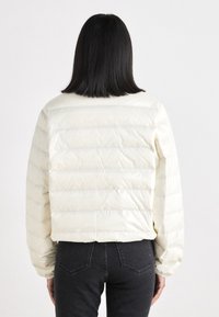 Elfenben pufferjakke med quiltet design, med en kortet snitt, elastiske mansjetter og en glatt, skinnende tekstur. Vist bakfra.