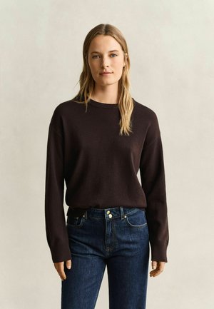 Jonge vrouw met lang blond haar, gekleed in een donkerbruine lange mouwen trui en blauwe spijkerbroek, staand voor een eenvoudige lichte achtergrond.