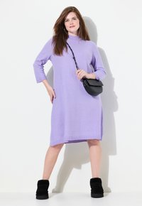 Lavendelfarbenes Strickkleid mit hohem Kragen, drei Viertelärmeln und geradem Schnitt. Kombiniert mit einer kleinen schwarzen Umhängetasche und schwarzen Stiefeletten.