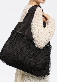 Czarna torba typu tote z siatkowym wzorem i solidnymi tkaninowymi akcentami. Charakteryzuje się gładką teksturą oraz prostokątną naszywką z logo na boku.