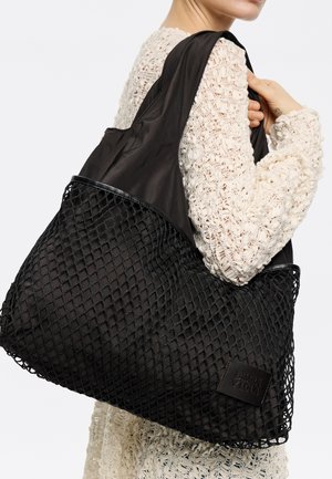 Czarna torba typu tote z siatkowym wzorem i solidnymi tkaninowymi akcentami. Charakteryzuje się gładką teksturą oraz prostokątną naszywką z logo na boku.