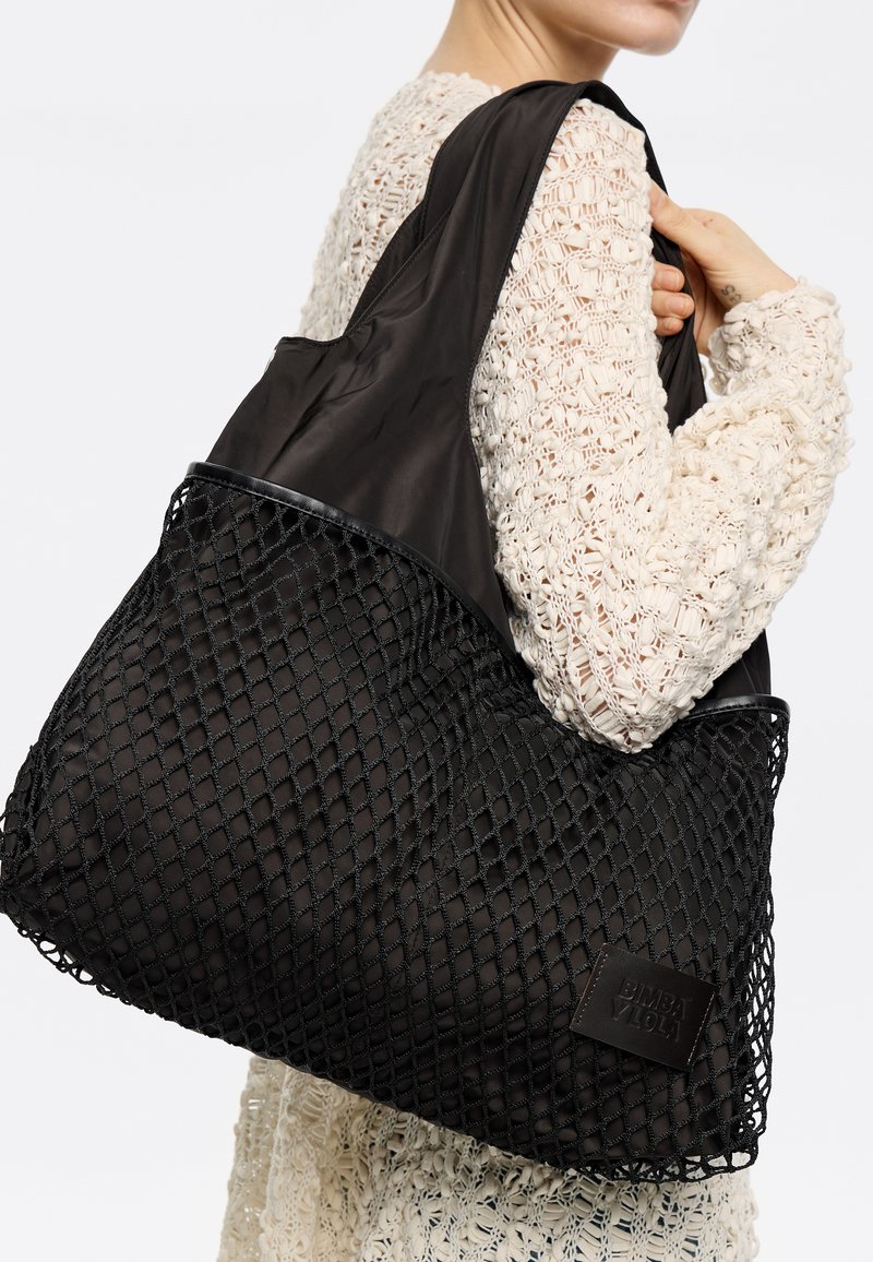 Czarna torba typu tote z siatkowym wzorem i solidnymi tkaninowymi akcentami. Charakteryzuje się gładką teksturą oraz prostokątną naszywką z logo na boku.