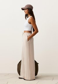 Woman in beige wide-leg pants, white sleeveless crop top, brown cap, standing in profile with hands in pockets against plain background.