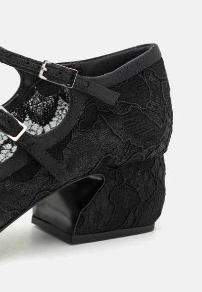 Black lace Mary Jane shoe with a low block heel, featuring adjustable straps and textured fabric with floral patterns.