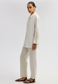 Camicia color crema a maniche lunghe con chiusura a bottoni e polsini arrotolati, abbinata a pantaloni su misura dello stesso colore e sandali neri con cinturini.