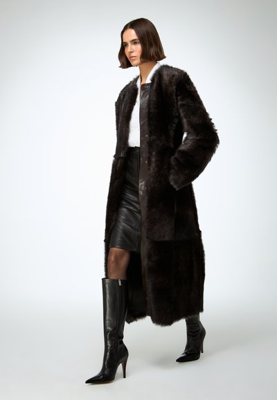 Long manteau en fausse fourrure marron avec une texture lisse, doté d'une fermeture à boutons sur le devant et de poches latérales, porté sur une jupe en cuir noire et des bottes montantes jusqu'au genou.