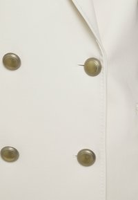 Witte stoffen blazer met een dubbelgeknoopt ontwerp, voorzien van twee ronde metallic knopen met een gestructureerde rand en een gladde afwerking.