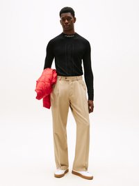 Haut à manches longues noir avec logo, pantalon ample beige et une veste matelassée rouge drapée sur le bras. Baskets blanches avec semelles brunes.