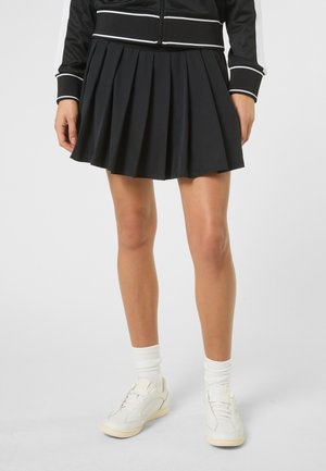 Vrouw die een zwarte geplooide rok, een zwart jack met witte streepdetails, witte sportsokken en witte sneakers draagt, staand op een witte achtergrond.