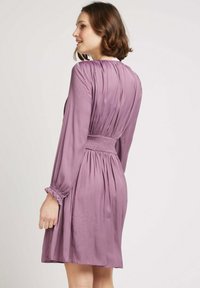 Robe lavande à manches longues, taille smockée et poignets volants. Texture lisse avec des plis le long du dos et au-dessus des épaules.