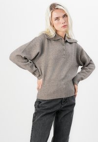 Jasnobrązowy sweter z dzianiny z kołnierzykiem, plisą na guziki, ściągaczami przy rękawach i u dołu. Noszony z ciemnoszarymi spodniami typu wide-leg.