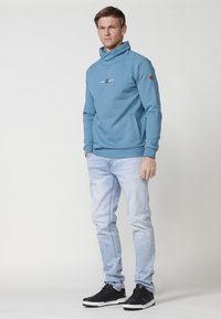 Felpa leggera blu con un alto colletto e stampa grafica sul petto, caratterizzata da maniche lunghe e tasche laterali. Indossata con jeans chiari e sneaker nere.