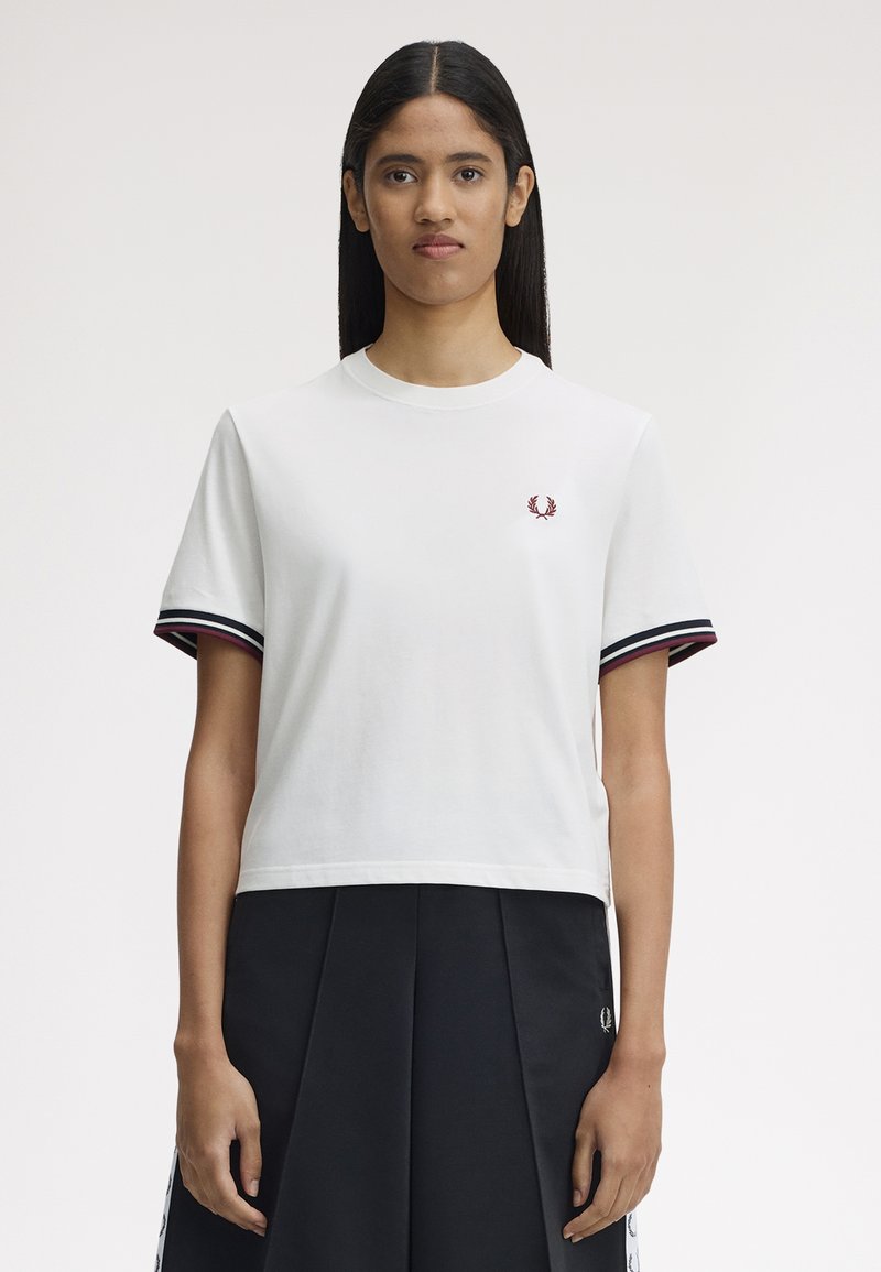 Jeune femme aux longs cheveux foncés portant un chemisier blanc à manches courtes avec des poignets rayés et une jupe plissée noire, sur un fond uni.