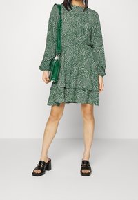 Vihreä kuvioitu mekko pitkähihaisilla, kerroksilla varustetuilla röyhelöillä, yhdistetty mustiin paksupohjaisiin sandaaleihin ja vihreään olkalaukkuun.