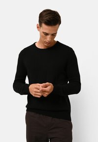 Zwart gebreid sweater met een ronde hals, geribde manchetten en zoom. Model draagt een paarse broek en heeft een neutrale uitdrukking.