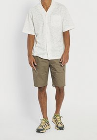 Wit overhemd met korte mouwen en patroon, olijfgroene cargobroekshort en beige sneakers met groene accenten en donkere strepen, die casual zomerse kleding laten zien.