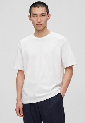 Jeune homme aux cheveux courts portant un t-shirt blanc uni et un pantalon foncé, debout avec une main dans la poche.