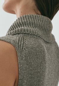 Maglione senza maniche in filato grigio, con un alto colletto a coste e motivi a maglia testurizzati. Il tessuto appare morbido e confortevole.