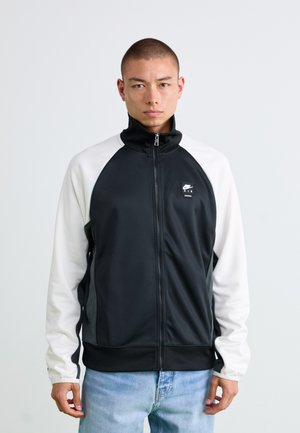 Giacca zip-up bianca e nera con colletto alto, maniche raglan e tasche frontali. Presenta il logo Nike Air sul lato sinistro del petto. Texture liscia.