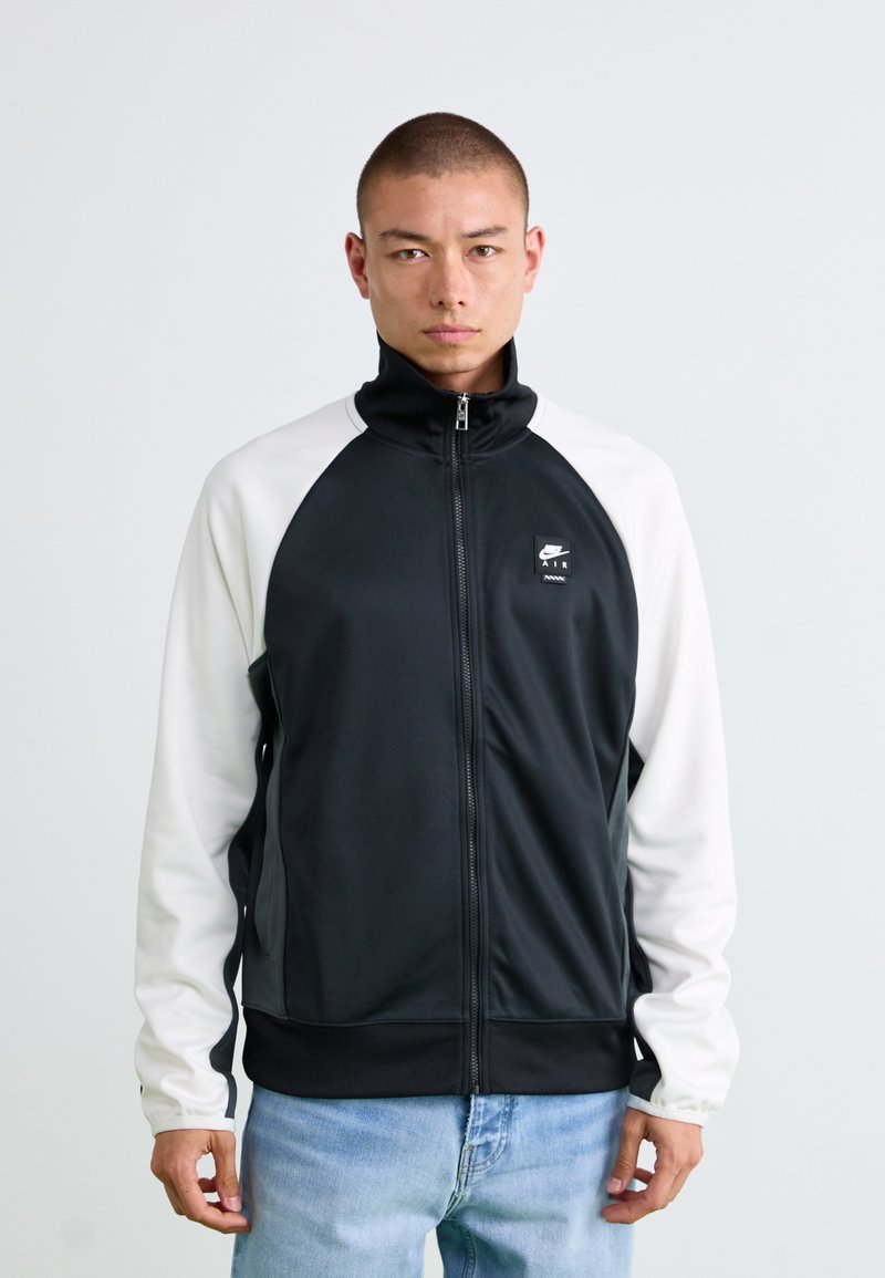 Crna i bijela zip-up jakna s visokim ovratnikom, raglan rukavima i prednjim džepovima. Sadrži Nike Air logo na lijevoj strani prsa. Glatka tekstura.