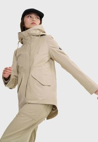 Beige wasserdichte Jacke mit Kapuze, Frontreißverschluss, zwei großen schrägen Taschen und verstellbaren Kordeln. Das Model trägt eine schwarze Mütze und posiert seitlich.