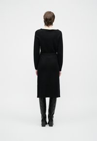 Person med kort hår som har på seg en svart langermet kjole og knelengde svarte støvler, står med ryggen til på en ensfarget bakgrunn.