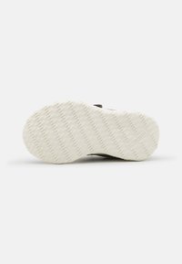 Semelle de chaussure avec motif de grip texturé blanc cassé et nom de marque « hummel » embossé près du talon, photographiée sur fond blanc.