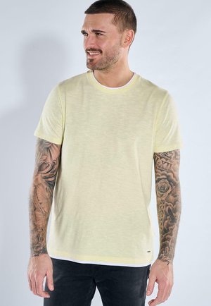 Man with short hair and tattoos on both arms wearing a light yellow short-sleeve T-shirt and black pants, looking to the side.