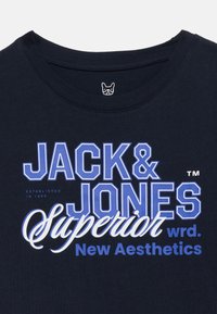 Γιακάς μπλε ναυτικού μπλουζάκι με το κείμενο «JACK & JONES Superior wrd. New Aesthetics» σε λευκό και μπλε χρώμα, και ένα μικρό λογότυπο προσώπου σκύλου μέσα στον γιακά.