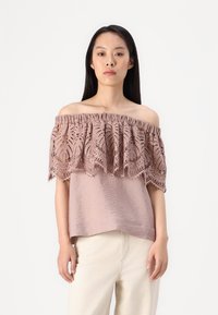 Top off-the-shoulder in rosa polveroso con intricate pizzo sovrapposto e orlo arricciato; tessuto leggero e testurizzato, abbinato a pantaloni chiari.