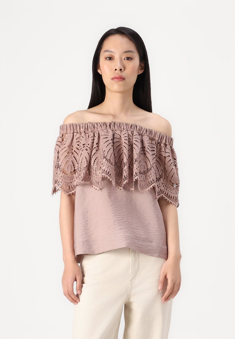 Top off-the-shoulder in rosa polveroso con intricate pizzo sovrapposto e orlo arricciato; tessuto leggero e testurizzato, abbinato a pantaloni chiari.