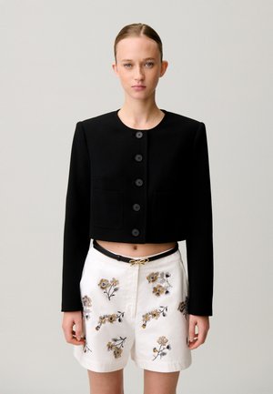 Femme aux cheveux plaqués en arrière portant une veste courte noire boutonnée et un short blanc brodé de fleurs avec une ceinture noire.