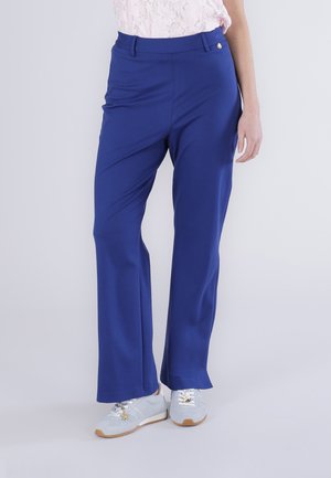 Kobieta w niebieskich prostych spodniach, białych trampkach z zawieszkami w kształcie gwiazdek oraz jasnoróżowej koronkowej bluzce, stojąca na tle o jednolitym kolorze.