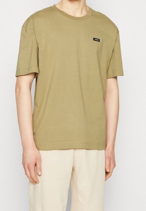 T-shirt in cotone verde oliva con maniche corte, scollatura rotonda e un piccolo patch logo nero sul petto. Texture liscia, vestibilità comoda.