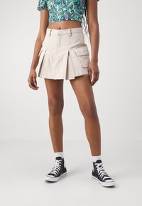Beige cargo szoknya redőkkel, oldalsó zsebekkel és testhezálló derékkal. Fekete magas szárú tornacipővel és fehér zoknival párosítva.