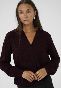 Blusa rosso scuro con un sottile motivo a zebra nera, scollatura a V, maniche lunghe e polsini arricciati. Indossata con anelli d'argento sulle mani.