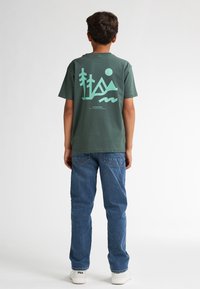 Groene katoenen T-shirt met een grote turquoise graphic van bomen en bergen op de rug, gecombineerd met blauwe denim jeans en witte schoenen.