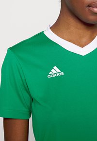 Zelené športové tričko s bielym V-výstrihom. Obsahuje vyšívané biele logo Adidas na ľavej strane. Vyrobené z hladkej látky.