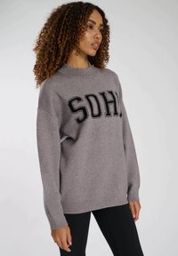 Grå stickad tröja med rund hals och stora bokstäver "SOHO" i svart. Lös passform med långa ärmar och ribbade muddar.