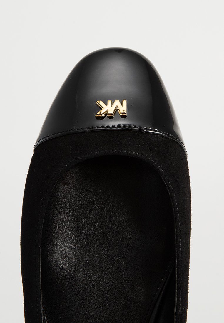 Zapato plano negro con puntera de charol brillante y cuerpo de ante cosido, con un detalle de emblema dorado en la parte delantera.
