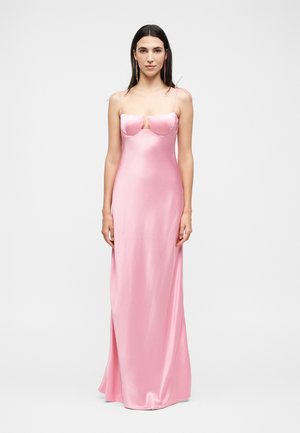 Γυναίκα με μακριά σκούρα μαλλιά, που φοράει ένα μακρύ μέχρι το πάτωμα, αμάνικο ανοιχτό ροζ σατέν φόρεμα με δομημένο σουτιέν και λεπτές τιράντες.
