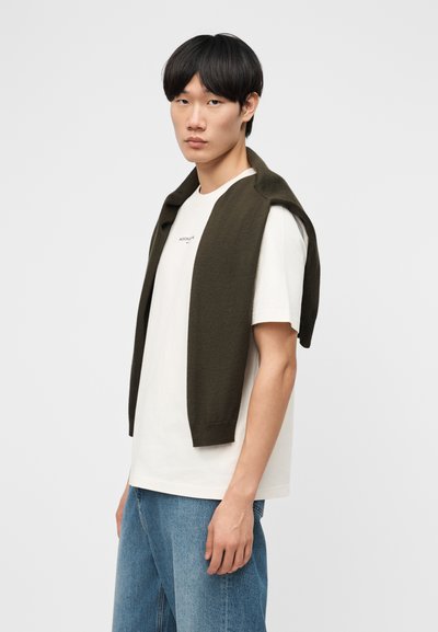 Νέος άνδρας με μαύρα μαλλιά, φορώντας λευκό μπλουζάκι, μπλε τζιν και σκούρο πράσινο πουλόβερ ριγμένο στους ώμους, στεκόμενος μπροστά σε φωτεινό φόντο.