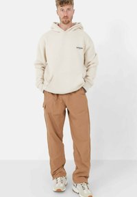 Beige hoodie met een voorvak, gedragen met bruine cargobroek en meerkleurige sneakers, staande tegen een effen witte achtergrond.